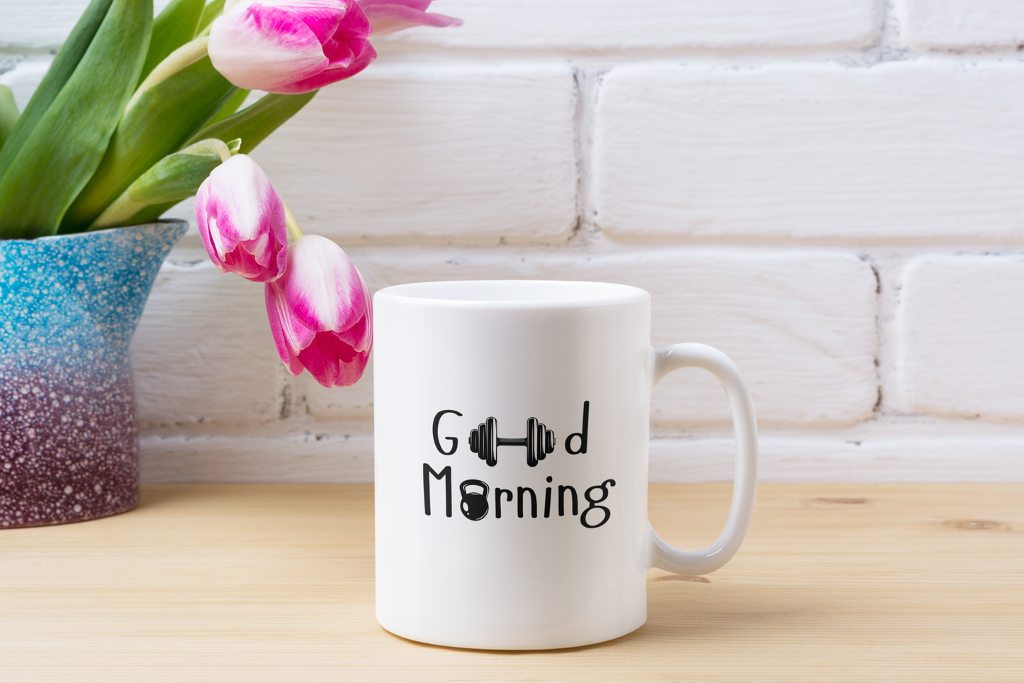 “Good Morning” ceramic mug on a wooden table with pink flowers and a book – calm and relaxing morning setup by FitBOUND