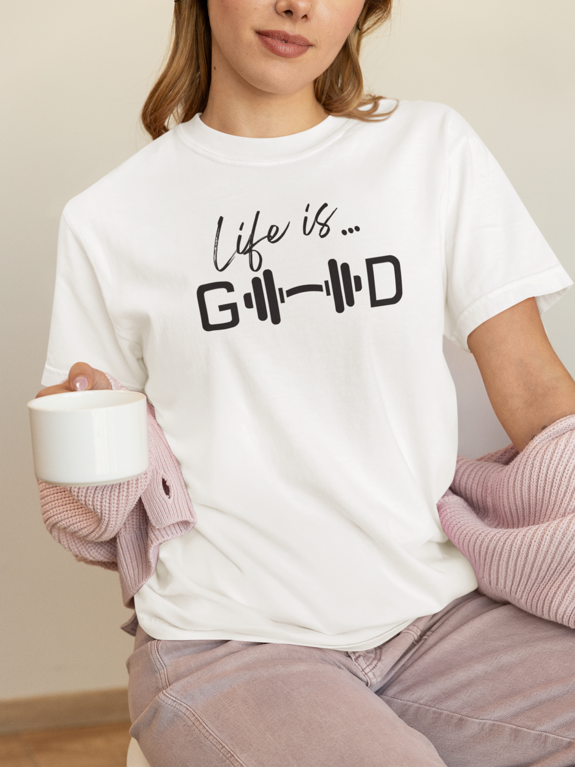 Woman wearing white motivational t-shirt with barbell design and text “Life is Good,” casual fitness style.