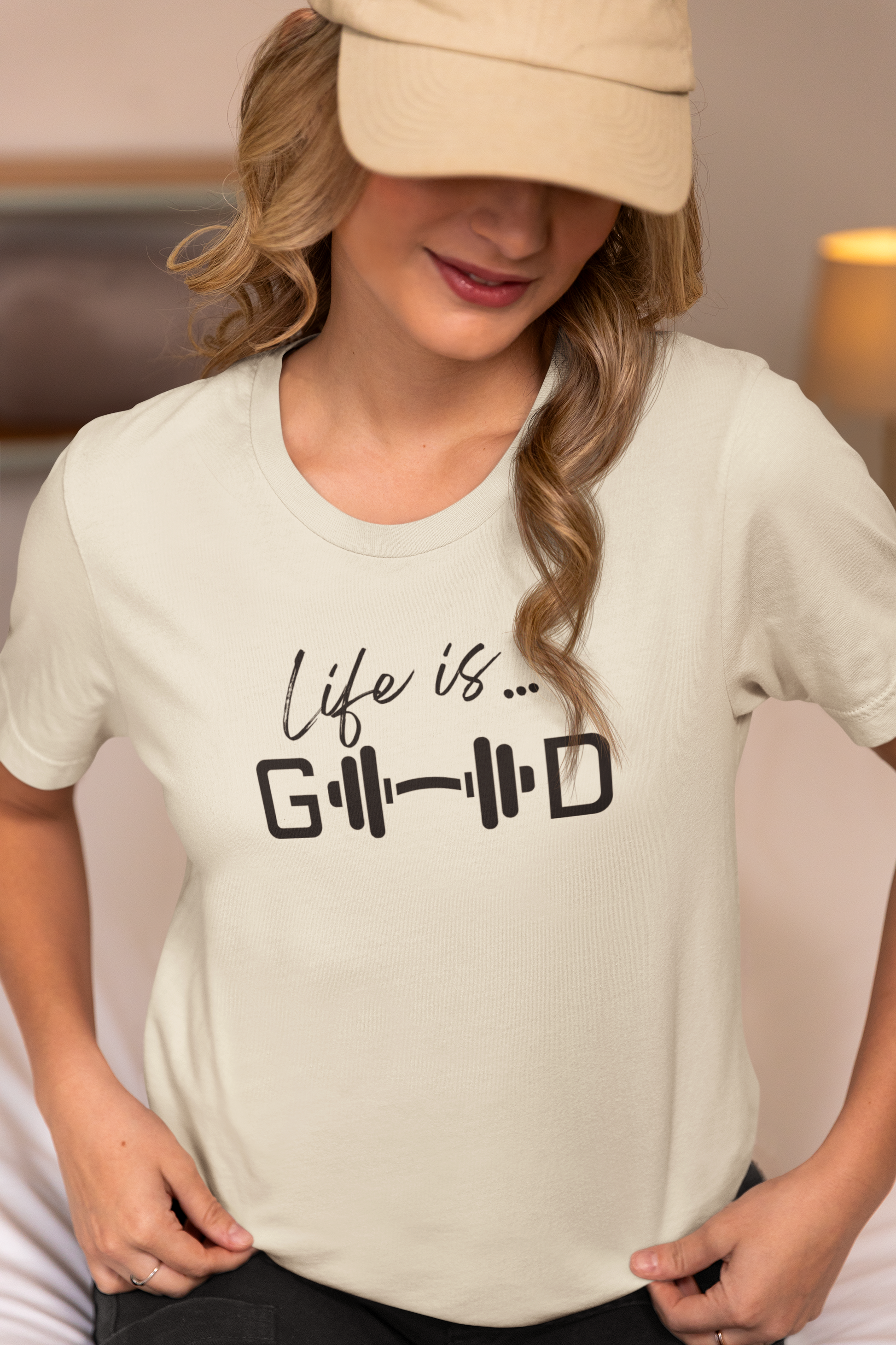 Woman wearing beige workout t-shirt with text “Life is Good” and barbell graphic, lifestyle outfit.